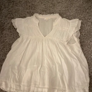 Vit blus med volang från Gina Tricot - Supersöt vit blus från Gina Tricot i luftig bomull. Blusen har v-ringning, volangdetaljer vid ärmarna och lätt rynkade partier upptill. 