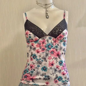 Blommig linnetopp med spetsdetalj - Supersöt linnetopp med blommigt mönster i rosa, turkos och grå toner på vit botten. Toppen har svarta spetsdetaljer vid bysten och tunna axelband. Perfekt för dig som vill ha en feminin och fräsch look till sommaren.