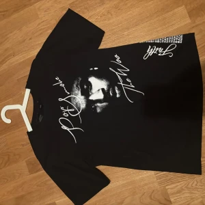 Pop Smoke svart t-shirt med tryck - Svart t-shirt med stort vitt tryck av Pop Smoke och texten 'The Woo' på framsidan. Baksidan har ett coolt V-format röktryck och låtlista i vitt. Rund hals och korta ärmar, tillverkad i mjuk bomull. Perfekt för dig som gillar streetwear och hiphop. (Liten passform)