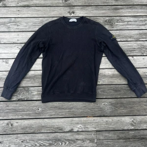 Svart sweatshirt från Stone Island - Säljer en svart långärmad sweatshirt från Stone Island med klassisk rund hals och ikonisk patch på ärmen. Tröjan är i bomull och har en clean, stilren look som passar perfekt till streetwear. Ribbstickade muddar vid ärmslut och nederkant. Nu pris 2400kr köp för ca 1 år sedan 