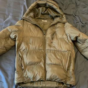 Olivgrön pufferjacka med huva Kappa - Säljer en olivgrön pufferjacka med huva och dragkedja framtill. Jackan har två sidofickor med dragkedja och en bröstficka med dragkedja. Perfekt för kalla dagar och riktigt skön att bära tack vare det vadderade materialet.