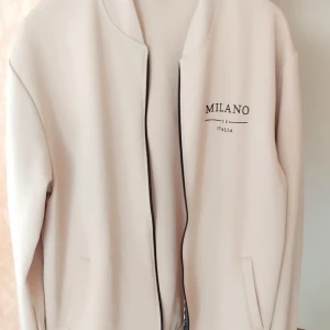Unik Beige ziptröja med text Milano Italia - Snygg beige ziptröja med text Milano Italia med svart dragkedja och tryck både fram och bak. Jackan har ribbade muddar, två fickor och klassisk ziptröja. Perfekt för dig som gillar stilren och enkel design.