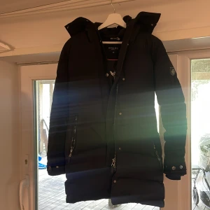 Svart Hollies Jacka st 36/S - Säljer en svart lång dunjacka från Hollies i storlek 36. Jackan har huva, två dragkedjefickor och snygga detaljer med knappar vid ärmslut. Perfekt för kalla dagar och har en stilren look som funkar till det mesta.