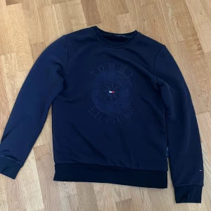 Mörkblå sweatshirt Tommy Hilfiger - Snygg mörkblå sweatshirt från Tommy Hilfiger med rund hals och stort broderat logomärke på bröstet. Tröjan har långa ärmar och ribbade muddar vid hals, ärmslut och nederkant. 