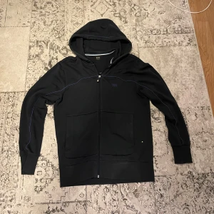 Mörkblå  hoodie från Hugo Boss med dragkedja - Mörkblå hoodie från Hugo Boss med blå detaljer och broderad logga på bröstet. Tröjan har huva, två stora fickor framtill och hel dragkedja. Perfekt för dig som gillar stilren och sportig look.