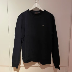 Marinblå sweatshirt från Morris - En stilren marinblå sweatshirt från Morris med rund hals och liten broderad logga på bröstet. Tröjan har långa ärmar och ribbade muddar vid ärmslut och nederkant. Perfekt för en clean och enkel look. Den är i extremt bra skick då den endast har använts 2 gånger. 