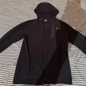 Svart vindjacka från Under Armour - Svart vindjacka från Under Armour med huva och dragkedja. Jackan har en bröstficka med dragkedja och diskret logga på bröstet. Långärmad modell med sportig känsla, perfekt för blåsiga dagar. Snygga röda detaljer vid huvan. Dragkedjan är trasslig den går att stänga men åker upp ofta 