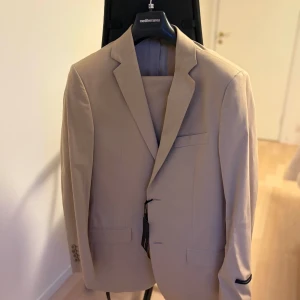 Beige kostym – Mediterranea, ny med tagg (stl 50) - Säljer nu en beige kostym. Väldigt snygg passform och modern snitt. Kavajen har två framfickor med lock, bröstficka samt innerfickor. Mjukt tyg och bra kvalitet – redo att användas direkt.  Märke: Mediterranea  Storlek: 50  Färg: Beige  Skick: Helt ny, aldrig använd  Tag kvar: Ja  Nypris: 4195 kr   Foder: Matchande ljusfärgat foder  Byxor: Matchande, med pressveck och bakficka med knapp