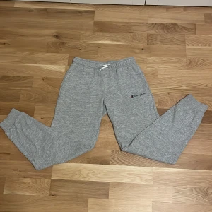 Grå mjukisbyxor från Champion - Gråa mjukisbyxor från Champion med elastisk midja och dragsko. Byxorna har en broderad Champion-logga på ena benet och muddar vid bensluten. Perfekta för chill dagar eller träning. Tillverkade i mjukt material för extra komfort.