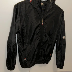 Svart vindjacka från Moncler med huva - Svart vindjacka från Moncler med huva och dragkedja framtill. Jackan har elastiska muddar, diskreta röda och vita detaljer vid huva och fickor samt Moncler loggan på armen. Väldigt fin om du vill se extra fräsch ut när du går ut med kompisar eller vill sticka ut i skolan!!