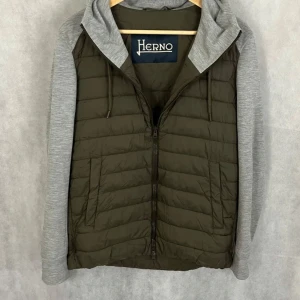 Grön pufferjacka med grå ärmar från Herno - Snygg pufferjacka från Herno i olivgrönt med quiltad kropp och grå långärmade ärmar i mjukt material. Jackan har huva med snörning och dragkedja framtill. Perfekt för dig som gillar en sportig och chill grisch stil. S/M nypris 5000kr