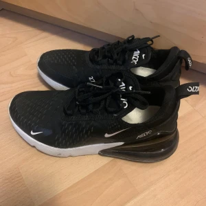 Nike Air Max 270 i svart, strl 38 - Säljer ett par svarta Nike Air Max 270 sneakers med vit sula och tydlig Air Max-bubbla i hälen. Skorna har mesh-ovandel, snörning och diskreta vita loggor. Perfekta för dig som gillar sportig och modern stil.