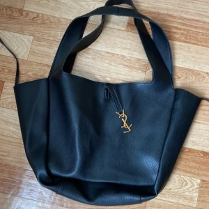 Svart axelväska från Saint Laurent - Stilren svart axelväska från Saint Laurent i skinn med guldigt YSL-hänge framtill. Väskan har breda axelband och rymligt huvudfack med innerficka med dragkedja. Perfekt för dig som vill ha en lyxig och praktisk väska till vardagen.
