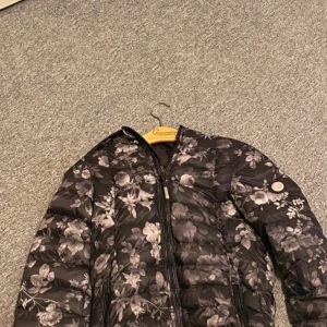 Blommig svart pufferjacka från Amisu - Snygg svart pufferjacka från Amisu med blommigt mönster i grå toner. Jackan har huva, dragkedja framtill och två fickor med dragkedja. Perfekt för dig som vill sticka ut lite extra under kyliga dagar.Jackan är använd en gång och den ser helt ny ut . pris går att diskutera. 