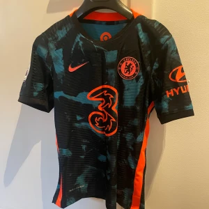 Chelsea FC kortärmad matchtröja Nike - Chelsea FC matchtröja från Nike med coolt svart och blått mönster, orange detaljer och loggor. Kortärmad med sportig passform, tillverkad i lätt och ventilerande material. Snygga sponsortryck på bröst och ärmar, perfekt för dig som älskar fotboll och vill sticka ut.