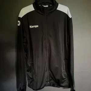 Svart träningsjacka från Kempa XL - Sportig svart träningsjacka från Kempa i storlek XL med vita detaljer på axlarna. Jackan har hel dragkedja, höga krage och två fickor med dragkedja. Perfekt för träning eller uppvärmning. Materialet är lätt och känns svalt mot huden. Tröjan är oanvänd då jag beställde fel storlek.