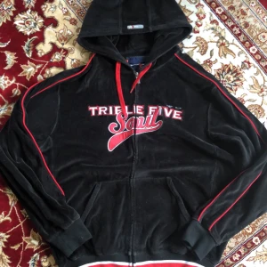 Svart velour hoodie Triple Five Soul - Svart hoodie i mjuk velour från Triple Five Soul med röda detaljer och broderad logga framtill. Tröjan har huva, dragkedja och fickor samt röda ränder längs ärmarna och ribbade muddar med röd-vit rand vid midjan. Kan mötas i sthlm.