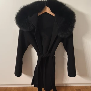 Svart kappa med äktapäls och bälte - Snygg svart kappa med stor huva kantad av fluffig fuskpäls. Jackan har knytskärp i midjan för en schysst siluett och långa ärmar. Perfekt för kalla dagar när du vill se extra stylish ut.
