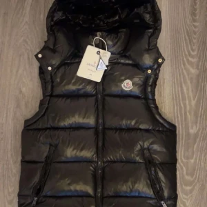 Svart dunväst från Moncler med huva - Snygg svart dunväst från Moncler med glansig finish och avtagbar huva. Västen har två dragkedjeförsedda fickor, Moncler-logga på bröstet och quiltad design. Perfekt för lager-på-lager och streetstyle.