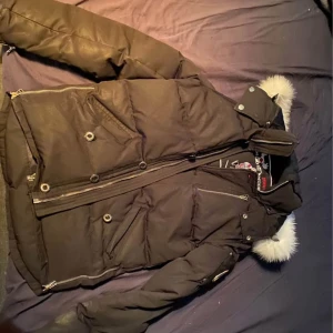 Svart dunjacka med päls från Moose Knuckles - Snygg svart dunjacka från Moose Knuckles med fluffig vit päls på huvan. Jackan har flera dragkedjor, knappar och coola detaljer i metall. Perfekt för kalla dagar och har en riktigt urban vibe. Flera fickor och en stilren look som passar streetwear.