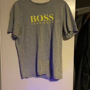 Grå t-shirt från Hugo Boss - Snygg grå t-shirt från Hugo Boss med gul logga tryckt på framsidan. 1 litet hål i varje armhåla och loggan har spruckit på ett ställe 