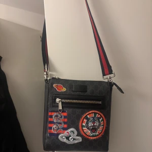 Gucci axelväska med patches och GG-mönster - Säljer en svart axelväska från Gucci med klassiskt GG-monogram och coola broderade patches framtill, bland annat en tiger och planet. Väskan har en bred röd- och blårandig axelrem och dragkedja framtill. Materialet är skinn och väskan har en fyrkantig form. Såklart Äkta orginall kan mötas upp i malmö
