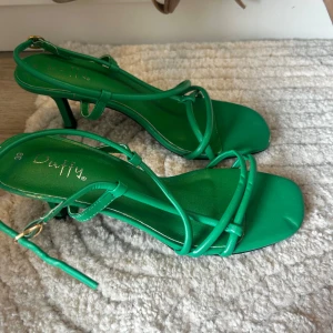 Gröna sandaler med klack från Duffy - Snygga gröna sandaler från Duffy med smala remmar och fyrkantig tå. Skorna har en elegant klack och justerbart spänne vid ankeln. Perfekta för att lyfta din outfit med en pop av färg.
