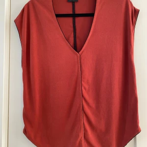 Rostfärgad topp från By Malene Birger - Snygg rostfärgad topp från By Malene Birger med djup v-ringning och minimalistisk design. Toppen är tillverkad i mjuk polyesterblandning med elastan och har svarta detaljer längs kanterna. .