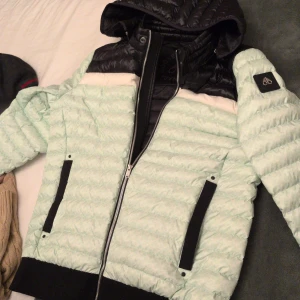 Moose Knuckles dunjacka - mintgrön & svart - Snygg pufferjacka från Moose Knuckles mintgrön med svarta detaljer och huva. Jackan har dragkedja framtill, svarta muddar och två fickor med svarta band. Perfekt för kyliga dagar och riktigt street. Strl M