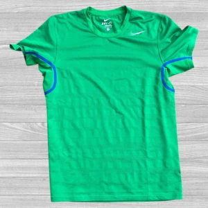 Grön Nike Dri-Fit t-shirt - Snygg grön t-shirt från Nike med Dri-Fit teknologi, perfekt för träning. T-shirten har korta ärmar, rund hals och diskreta mesh-paneler för extra ventilation. Blå detaljer vid ärmarna och klassisk Nike-logga på bröstet. Tillverkad i lätt polyester.