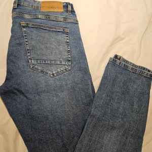 Blå jeans från SMOG, rak modell - Klassiska blå jeans från SMOG i rak passform. Jeansen har fem fickor, snygga slitningar och kontrastsömmar. Tillverkade i bomull med en autentisk denimkänsla. Perfekta för en avslappnad och stilren look.