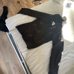 Svart Sail Racing GoreTex jacka - Svart jacka från Sail Racing i Gore-Tex-material med hög krage och flera dragkedjeförsedda fickor. Jackan har logga på bröstet och ärmen samt justerbar nederkant med snörning. Perfekt för dig som gillar stilren och funktionell design. Ny