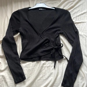 Svart omlottopp XS - Svart omlottopp i storlek XS. Toppen har långa ärmar, croppad passform och knyt i sidan. V-ringad halsringning och mjukt, tunt material som känns skönt mot huden. Perfekt att styla med jeans eller kjol 🖤