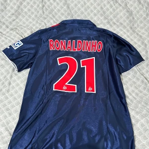 PSG Ronaldinho 21 Nike fotbollströja - Säljer en klassisk Paris Saint-Germain fotbollströja med Ronaldinho och nummer 21 på ryggen. Tröjan är marinblå med en röd vertikal rand, vita detaljer och PSG-logga på bröstet. Nike Swoosh och Opel sponsortryck framtill. Tillverkad i glansigt polyester. Är i storlek M men passar L också. Pris kan diskuteras!