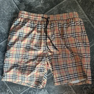 Rutiga Burberry shorts med snörning - Säljer dessa snygga shorts från Burberry i klassiskt beige rutigt mönster med svarta, vita och röda detaljer. Använda 1 gång så är inprincip nya i väldigt bra skick! Online kvitto finns också!