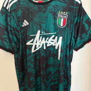 Italien x Stüssy x Adidas fotbollströja XL - Helt ny/oanvänd med taggar. Unik Italien fotbollströja i samarbete med Stüssy och Adidas. Tröjan har ett konstnärligt grafiskt mönster i mörkgrön och svart, med vita Stüssy-loggan på bröstet och Italiens klubbmärke med fyra stjärnor. Slim fit, kortärmad och tillverkad i lätt polyester. Storlek XL men kan passa som L. 