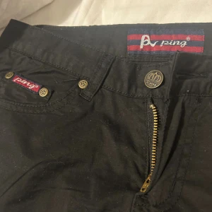Svarta byxor från Ping med raka ben - Svarta byxor från Ping med raka ben och klassisk passform. Byxorna har guldiga detaljer på knappar och dragkedja, samt broderad logga på fickan. Snygg midja och passar till många outfits. (Sitter löst på mig) 