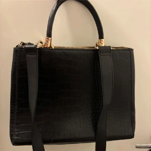 Svart handväska i croco-mönstrat skinn - Snygg svart handväska med croco-mönstrad yta och guldfärgade detaljer. Väskan har både handtag och axelrem, perfekt för att bära på olika sätt. Stängs med dragkedja upptill och har en stilren, fyrkantig form som ger en lyxig känsla.