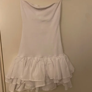 Vit kortklänning med volang - Supersöt vit kortklänning med flera lager volanger nedtill. Klänningen är ärmlös och har en enkel, rak passform upptill som övergår i en fluffig volangkjol. Prislappen är kvar!