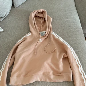 Beige croppad hoodie från Adidas - Säljer en snygg beige croppad hoodie från Adidas med klassiska vita ränder längs ärmarna och broderad logga på bröstet. Tröjan har huva med snörning. Perfekt för en chill och sportig look.