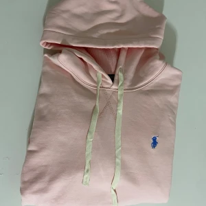 Ljusrosa hoodie från Polo Ralph Lauren - Säljer en ljusrosa hoodie från Polo Ralph Lauren med klassisk blå logga broderad på bröstet. Tröjan har huva med snörning, ribbade muddar och känguruficka. Perfekt för dig som gillar en clean och stilren look.