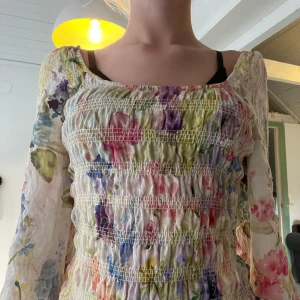 Blommig långärmad blus med öppen rygg - Superfin långärmad blus med blommigt mönster i pastelliga färger. Smockad överdel och ärmar, samt öppen rygg med knytband. Tunt, lätt material som känns luftigt. Utan lapp, men är väldigt fin och förgglad!