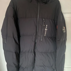 Svart dunjacka från Sail Racing M - Säljer en svart dunjacka från Sail Racing i storlek medium. Jackan har huva, dragkedjor framtill och på ärmen samt logga på bröstet och ärmen. Perfekt för kalla dagar och riktigt snygg streetstil.