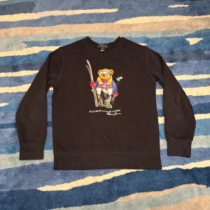 Svart sweatshirt Polo Bear Ralph Lauren - Svart sweatshirt från Polo Ralph Lauren med ikoniskt Polo Bear-tryck framtill. Tröjan har rund hals, långa ärmar och är gjord i väldigt mjuk bomull. Perfekt för dig som gillar streetstyle och vill sticka ut med en klassisk men lekfull look. OBS! Trycket på björnen har börjat spricka lite, men syns bara om man tittar nära, tips är att tvätta ut och in samt att tvätta så lite som möjligt då det är så trycket spricker. Pris kan diskuteras.
