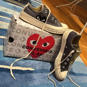 Converse x Comme des Garçons Play sneakers - Säljer ett par Converse x Comme des Garçons Play sneakers i mörkgrå canvas med den ikoniska svarta hjärtloggan på sidan. Skorna har vita gummisulor, rund tå och klassisk snörning. Perfekta för dig som vill ha en unik och stilren sneaker med streetkänsla. Snörade ur dem då jag håller på att rengöra snörena. Pris kan diskuteras.