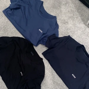 3-pack BOSS t-shirts blå & svart - Säljer tre stilrena t-shirts från BOSS i svart och två olika blå nyanser. Alla har rund halsringning och diskret BOSS-logga på bröstet. Perfekta för en clean och enkel look. Tillverkade i mjuk bomull och med kort ärm för maximal komfort.