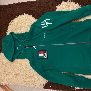 Grön hoodie Sail Racing ITA77 - Grön hoodie från Sail Racing med ITA77-tryck på ryggen och ärmen. Dragkedja framtill, huva med snörning och broderad patch på bröstet. Skön passform och perfekt för dig som gillar sportig stil.