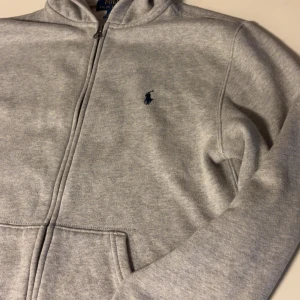 Ralph Lauren - Snygg ljusgrå hoodie från Polo Ralph Lauren med dragkedja och huva. Klassisk broderad logga i mörkblått på bröstet och stora fickor framtill. Pris inte hugget i sten. Storlek 14-16 