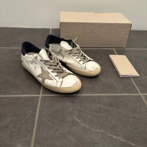 Golden Goose  - Golden Goose sneakers i vitt skinn med grå snörning och ikonisk grå stjärna på sidan. Skorna har beige sula och svart hälparti. Klassisk låg modell med rund tå och vintage-inspirerad look. Perfekta för dig som gillar streetstyle och unika detaljer.