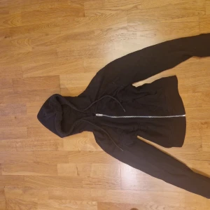 Svart hoodie med dragkedja - Svart hoodie med hel dragkedja framtill och snörning i huvan. Tröjan har långa ärmar och en klassisk, enkel design som passar till det mesta. Perfekt för chill dagar eller när du vill ha något bekvämt och stilrent.